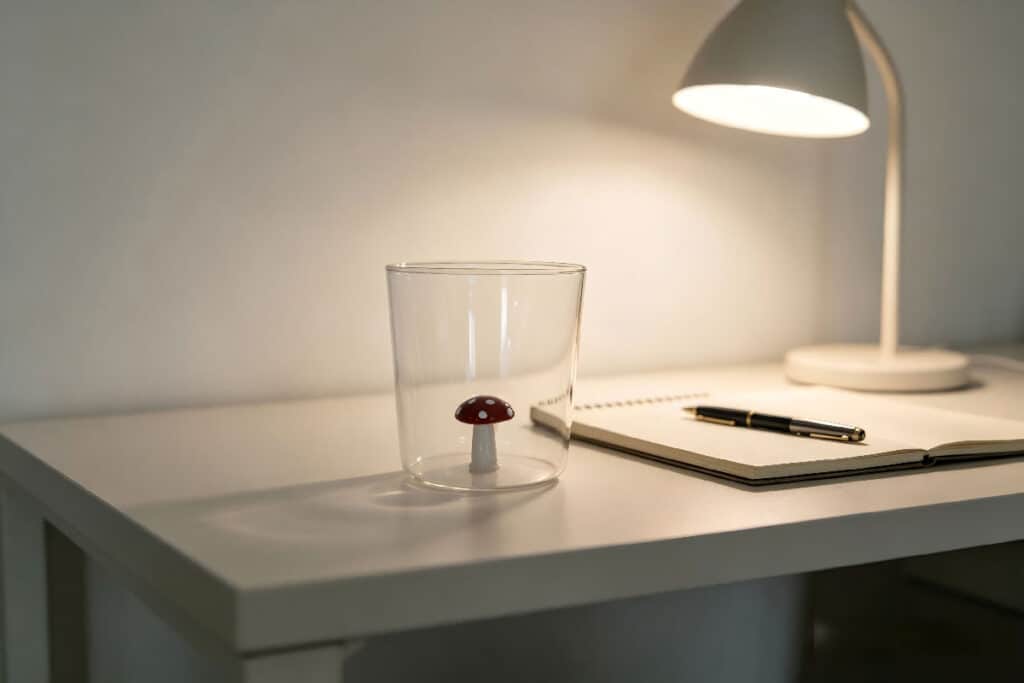 Clear straight glass tumbler with decorative red spotted mushroom inside, placed on minimalist white desk