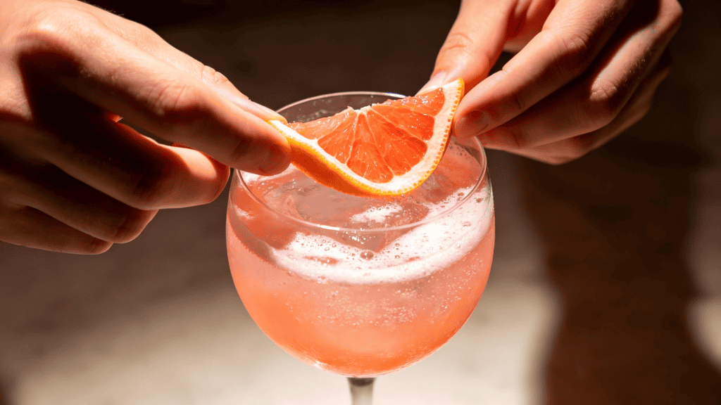 Clear stemmed cocktail glass with pink sparkling drink, garnished with a grapefruit slice being placed by hands