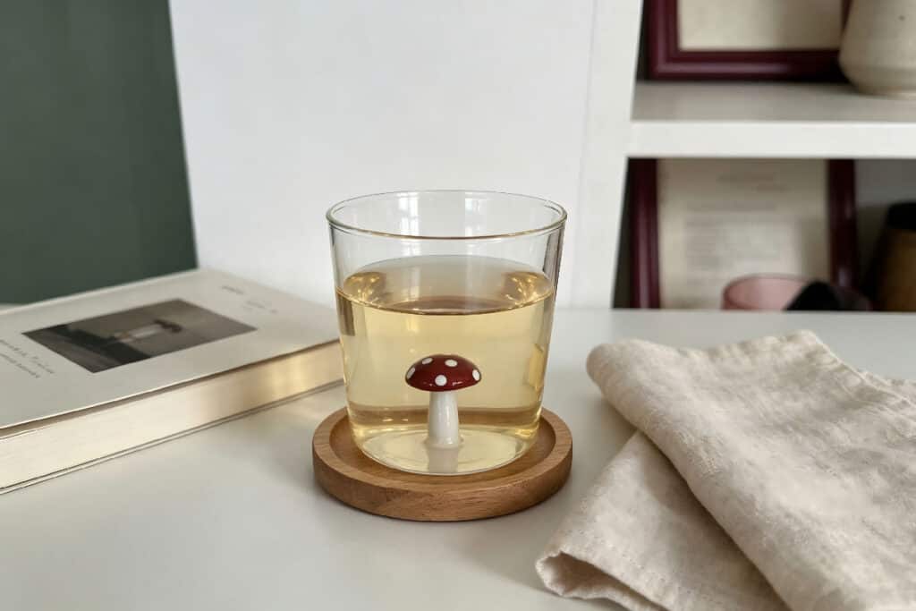 Clear glass tumbler with built-in red spotted mushroom ornament filled with tea, set on wooden coaster