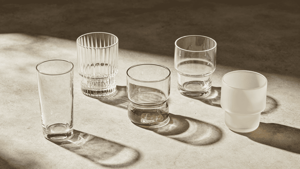 Set of five assorted clear and frosted glass tumblers, including ribbed, amber-tinted, and minimalist designs, displayed on a neutral sunlit surface