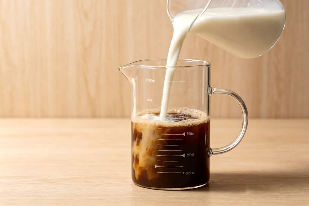 Glass measuring pitcher with measurement marks, pouring milk into coffee on wooden table