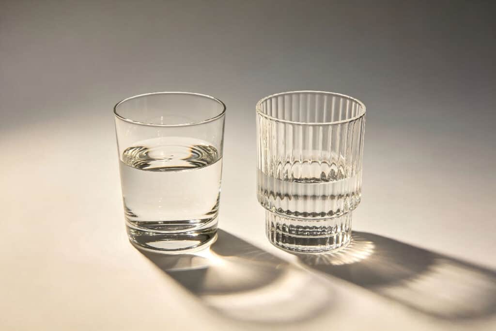 Two clear drinking glasses, smooth plain tumbler and vertical ribbed stacked glass with water on neutral background