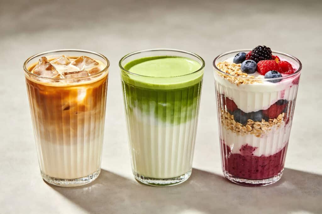 Three vertical ribbed glass tumblers filled with iced latte, matcha drink and berry yogurt parfait