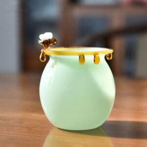 Handblown light green glass honey cup with glass bee and amber honey drip rim decor, placed on wooden table, lifestyle scene photo