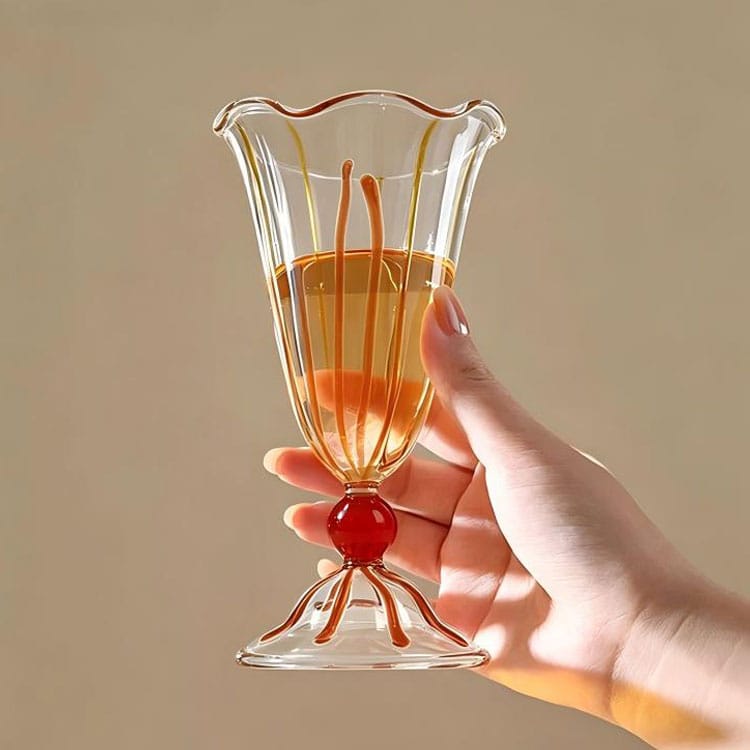 Handblown transparent stemmed glass cup with wavy rim, orange stripe decor, held in hand with golden beverage inside, for cocktail dessert milkshake