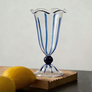 Handblown transparent stemmed glass cup with wavy rim, blue stripe decoration, placed on wooden board with lemons, for dessert, cocktail and milkshake