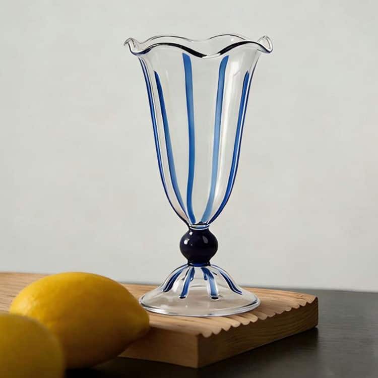 Handblown transparent stemmed glass cup with wavy rim, blue stripe decoration, placed on wooden board with lemons, for dessert, cocktail and milkshake